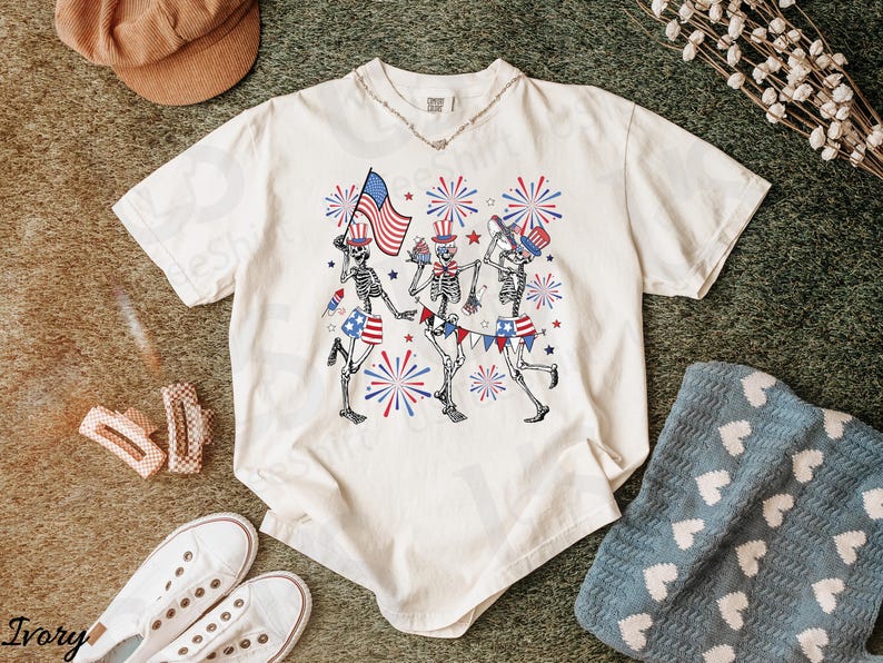 May include: Ivory-colored t-shirt featuring a festive design of dancing skeletons celebrating Independence Day. The skeletons are adorned with patriotic elements like the American flag and fireworks. The shirt is paired with white sneakers and accessories.