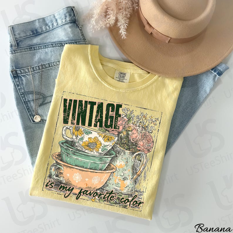 May include: A yellow t-shirt with the text "VINTAGE is my favorite color" and a graphic of teacups and flowers. The shirt is laid on top of blue jeans, with a necklace and a tan hat in the background.