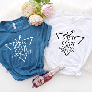 Op de afbeelding: Een blauw t-shirt met witte tekst Boats Booze & Bachelorette en een wit t-shirt met zwarte tekst Boats Booze & Bride, beide met een driehoek en anker afbeelding, vergezeld door een klein flesje Martini & Rossi mousserende wijn.