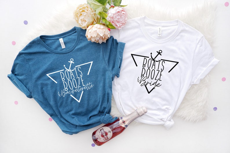Op de afbeelding: Een blauw t-shirt met witte tekst Boats Booze & Bachelorette en een wit t-shirt met zwarte tekst Boats Booze & Bride, beide met een driehoek en anker afbeelding, vergezeld door een klein flesje Martini & Rossi mousserende wijn.