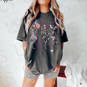 May include: Dark gray t-shirt featuring a graphic of dancing skeletons celebrating with American flags and fireworks. The skeletons wear patriotic hats and skirts. The text "USTeeShirt" is visible on the shirt.