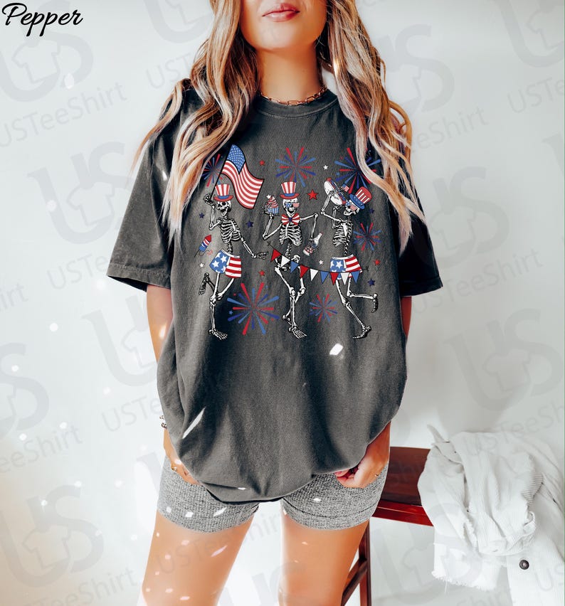 May include: Dark gray t-shirt featuring a graphic of dancing skeletons celebrating with American flags and fireworks. The skeletons wear patriotic hats and skirts. The text "USTeeShirt" is visible on the shirt.