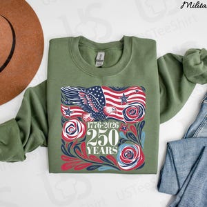 K&ouml;nnte beinhalten: Milit&auml;rgr&uuml;nes Sweatshirt mit einem grafischen Design mit einer amerikanischen Flagge, einem Adler und dem Text "1776-2026 250 YEARS". Das Design enth&auml;lt rote, wei&szlig;e und blaue Elemente. Ein brauner Hut und blaue Jeans sind ebenfalls im Bild.