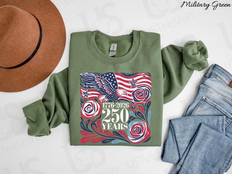 K&ouml;nnte beinhalten: Milit&auml;rgr&uuml;nes Sweatshirt mit einem grafischen Design mit einer amerikanischen Flagge, einem Adler und dem Text "1776-2026 250 YEARS". Das Design enth&auml;lt rote, wei&szlig;e und blaue Elemente. Ein brauner Hut und blaue Jeans sind ebenfalls im Bild.