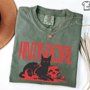 K&ouml;nnte beinhalten: Olivgr&uuml;nes T-Shirt mit dem Wort "ANTISOCIAL" in roten Buchstaben. Darunter befinden sich eine schwarze Katze, ein Totenkopf und ein Grabstein. Eine goldene Halskette liegt auf dem Shirt.