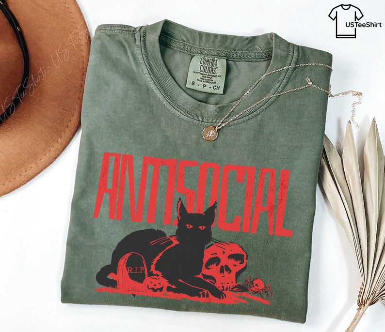 K&ouml;nnte beinhalten: Olivgr&uuml;nes T-Shirt mit dem Wort "ANTISOCIAL" in roten Buchstaben. Darunter befinden sich eine schwarze Katze, ein Totenkopf und ein Grabstein. Eine goldene Halskette liegt auf dem Shirt.