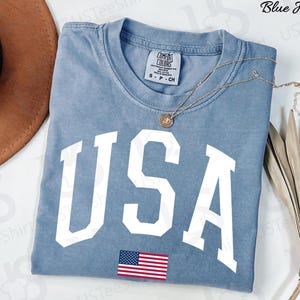 May include: A blue jean-coloured t-shirt with the word "USA" in large white block letters and a small American flag graphic. The shirt has a crew neck and short sleeves. A gold necklace with a pendant is draped across the shirt.
