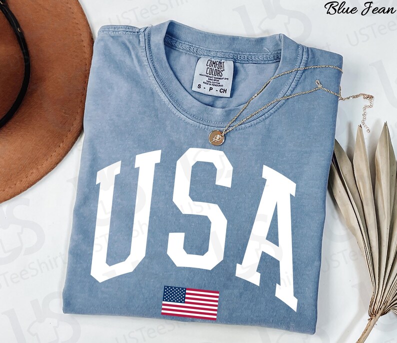 May include: A blue jean-coloured t-shirt with the word "USA" in large white block letters and a small American flag graphic. The shirt has a crew neck and short sleeves. A gold necklace with a pendant is draped across the shirt.