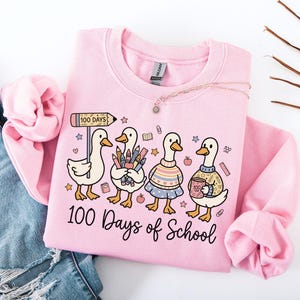 May include: Pink sweatshirt with a graphic of four cartoon ducks, each holding school supplies. The ducks are wearing sweaters and skirts. The text "100 Days of School" is printed below the ducks. A necklace is draped across the sweatshirt.