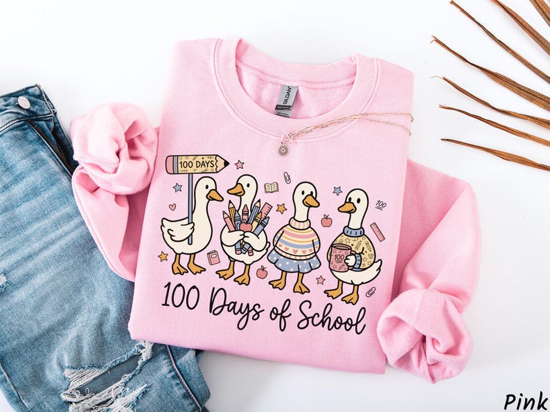 May include: Pink sweatshirt with a graphic of four cartoon ducks, each holding school supplies. The ducks are wearing sweaters and skirts. The text "100 Days of School" is printed below the ducks. A necklace is draped across the sweatshirt.