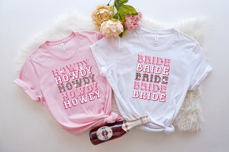 May include: Two t-shirts, one pink and one white, with text graphics. The pink shirt says "HOWDY" in a stacked design with a leopard print accent. The white shirt says "BRIDE" in a similar style. A bottle of sparkling wine is in the foreground.