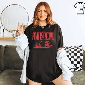 K&ouml;nnte beinhalten: Schwarzes T-Shirt mit dem Wort "ANTISOCIAL" in roter Schrift. Darunter befindet sich eine rote Grafik mit Totenk&ouml;pfen und anderen Elementen. Das T-Shirt wird getragen.
