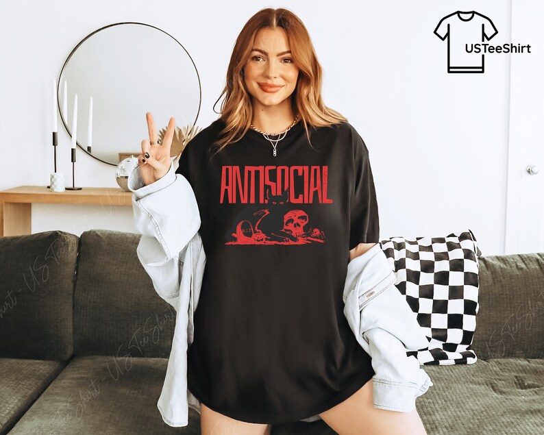 K&ouml;nnte beinhalten: Schwarzes T-Shirt mit dem Wort "ANTISOCIAL" in roter Schrift. Darunter befindet sich eine rote Grafik mit Totenk&ouml;pfen und anderen Elementen. Das T-Shirt wird getragen.