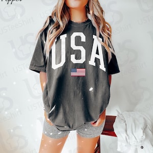 May include: Dark grey t-shirt featuring the word "USA" in large white letters and a small American flag graphic. A casual style t-shirt, suitable for everyday wear.