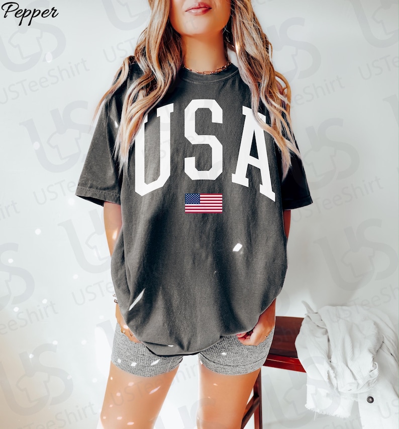 May include: Dark grey t-shirt featuring the word "USA" in large white letters and a small American flag graphic. A casual style t-shirt, suitable for everyday wear.