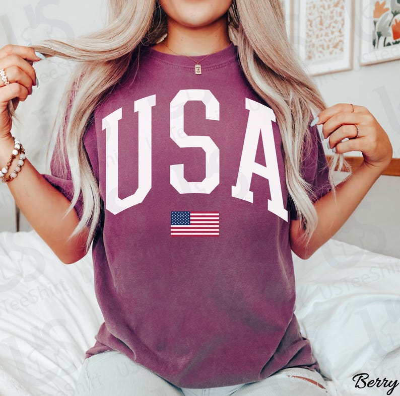 May include: Berry-coloured t-shirt featuring the word "USA" in large white block letters and a small American flag graphic. A casual style t-shirt, suitable for everyday wear.