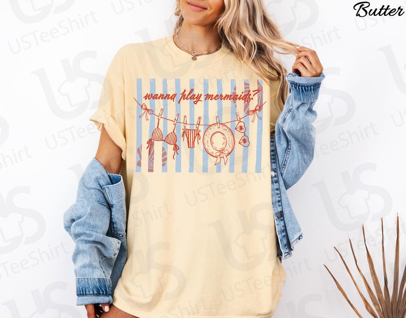 May include: A light yellow t-shirt with the text "wanna play mermaids?" above a graphic of a bikini, swimsuit, hat, and sunglasses hanging on a clothesline. The background features blue and white stripes. The person is wearing a denim jacket.