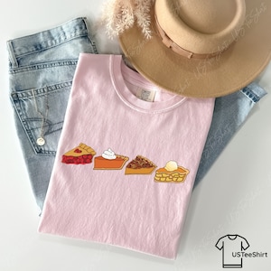 Puede incluir: Una camiseta rosa claro con un estampado gr&aacute;fico de cuatro porciones de tarta: cereza, calabaza con nata montada, nuez pecana y manzana con una bola de helado. La camiseta se presenta sobre vaqueros azules, un sombrero beige y un collar plateado.