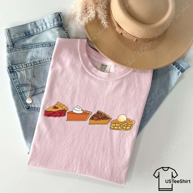 Puede incluir: Una camiseta rosa claro con un estampado gr&aacute;fico de cuatro porciones de tarta: cereza, calabaza con nata montada, nuez pecana y manzana con una bola de helado. La camiseta se presenta sobre vaqueros azules, un sombrero beige y un collar plateado.