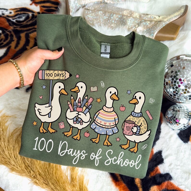 May include: Olive green sweatshirt with a graphic of four cartoon ducks celebrating 100 days of school. The ducks are holding school supplies and wearing sweaters. The text "100 Days of School" is written below the ducks.