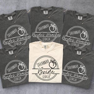 Op de afbeelding: Een verzameling van zes t-shirts, voornamelijk in antracietgrijs en één in crème. Elk shirt heeft een cirkelvormig ontwerp met de tekst "Savannah Brides Peaches Georgia" en een perzikafbeelding. Het crèmekleurige shirt zegt "Savannah Bride Georgia".