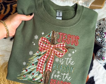 Könnte beinhalten: Ein olivgrünes Sweatshirt mit Rundhalsausschnitt und einem Grafikdruck eines Holzkreuzes, das mit einer rot-weiß karierten Schleife verziert ist. Ein stilisierter Weihnachtsbaum mit roten Kugeln und weißen Schneeflocken umgibt das Kreuz. Der Text lautet JESUS is the REASON for the SEASON.