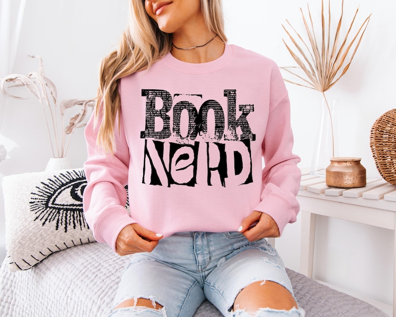 May include: A pale pink sweatshirt featuring the text "Book Nerd" in a black, blocky font. The long-sleeved top has a crew neck. The text is a prominent design element.