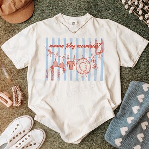 May include: Ivory-colored t-shirt with a graphic of a swimsuit, life preserver, and the text "wanna play mermaids?" on a blue and white striped background. The shirt is paired with white sneakers, hair clips, and a brown cap.