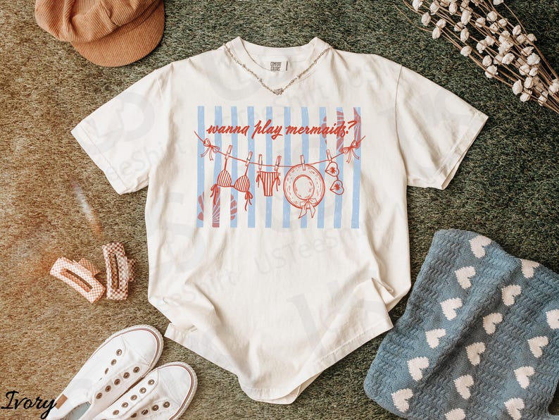 May include: Ivory-colored t-shirt with a graphic of a swimsuit, life preserver, and the text "wanna play mermaids?" on a blue and white striped background. The shirt is paired with white sneakers, hair clips, and a brown cap.