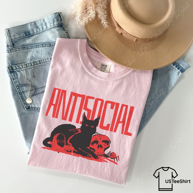 K&ouml;nnte beinhalten: Hellrosa T-Shirt mit dem Wort "ANTISOCIAL" in roter Schrift. Darunter ein schwarzer Kater, ein Totenkopf und ein Grabstein-Design. Das Shirt wird mit blauen Jeans und einem beigen Hut kombiniert.