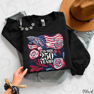 K&ouml;nnte beinhalten: Schwarzes Sweatshirt mit einem patriotischen Design mit einer amerikanischen Flagge, einem Adler und Rosen. Die Grafik enth&auml;lt den Text "1776-2026 250 YEARS". Das Sweatshirt wird mit einem Hut und Jeans gezeigt.