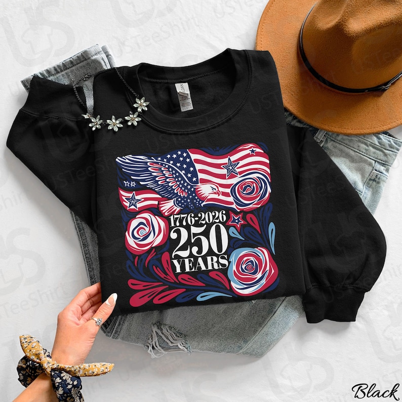 K&ouml;nnte beinhalten: Schwarzes Sweatshirt mit einem patriotischen Design mit einer amerikanischen Flagge, einem Adler und Rosen. Die Grafik enth&auml;lt den Text "1776-2026 250 YEARS". Das Sweatshirt wird mit einem Hut und Jeans gezeigt.