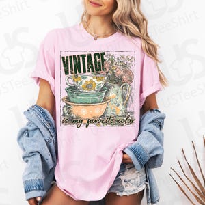 May include: Pink t-shirt with a graphic featuring stacked teacups, a pitcher of flowers, and the text "VINTAGE is my favorite color." The design has a vintage aesthetic.