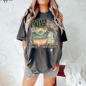 May include: Dark gray t-shirt with a vintage-style graphic. The design features stacked teacups and a floral pitcher, with the word "VINTAGE" above and "is my favorite color" below. The shirt is worn by a person.