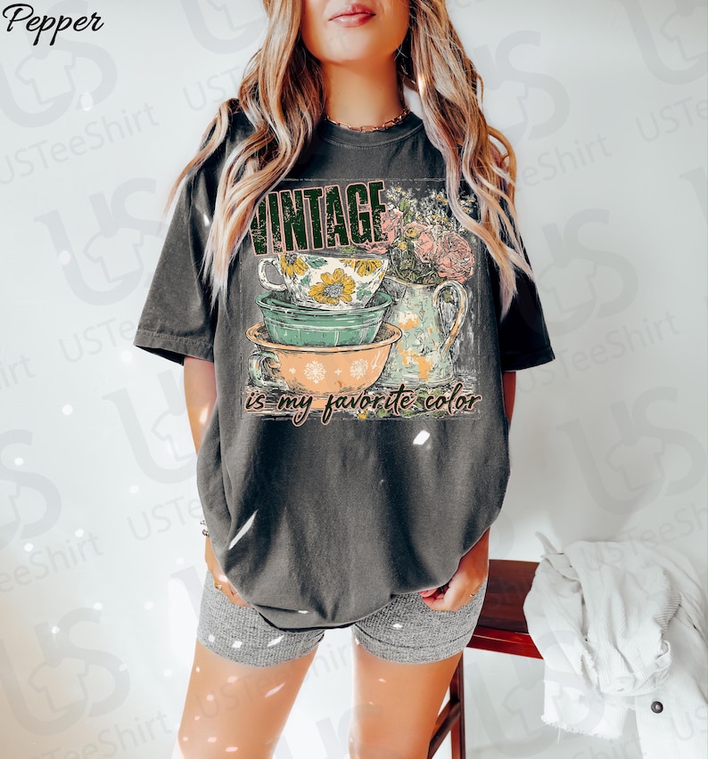 May include: Dark gray t-shirt with a vintage-style graphic. The design features stacked teacups and a floral pitcher, with the word "VINTAGE" above and "is my favorite color" below. The shirt is worn by a person.
