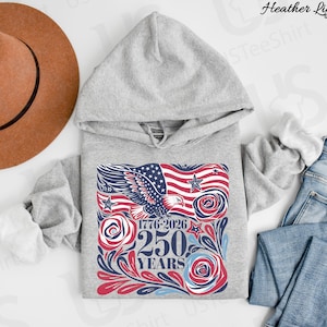 K&ouml;nnte beinhalten: Hellgrauer Kapuzenpullover mit einem grafischen Design mit einer amerikanischen Flagge, einem Adler und dem Text "1776-2026 250 YEARS". Das Design verwendet die Farben Rot, Wei&szlig; und Blau. Ein brauner Hut und blaue Jeans sind ebenfalls im Bild.