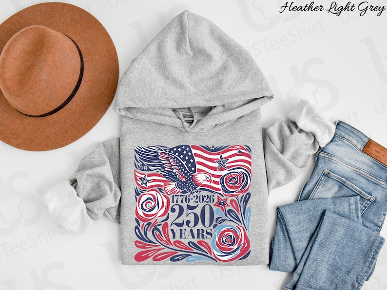 K&ouml;nnte beinhalten: Hellgrauer Kapuzenpullover mit einem grafischen Design mit einer amerikanischen Flagge, einem Adler und dem Text "1776-2026 250 YEARS". Das Design verwendet die Farben Rot, Wei&szlig; und Blau. Ein brauner Hut und blaue Jeans sind ebenfalls im Bild.