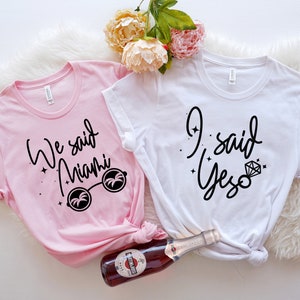 May include: Two t-shirts, one pink and one white, with black script. The pink shirt says "We said Miami" with sunglasses. The white shirt says "I said Yes" with a diamond ring graphic. A bottle of wine is in the image.