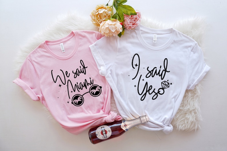 May include: Two t-shirts, one pink and one white, with black script. The pink shirt says "We said Miami" with sunglasses. The white shirt says "I said Yes" with a diamond ring graphic. A bottle of wine is in the image.