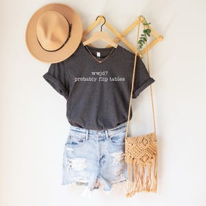May include: A dark gray t-shirt with the text "wwjd? probably flip tables" is paired with light wash denim shorts. A tan hat and a macrame crossbody bag complete the casual outfit. A gold necklace is visible.