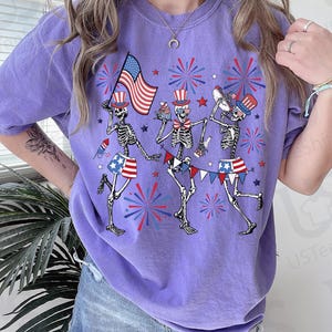 May include: A purple t-shirt featuring a festive design with three skeleton figures celebrating. They are adorned with patriotic hats and shorts, holding an American flag, cupcakes, and musical instruments. Fireworks and star accents complete the Fourth of July theme.