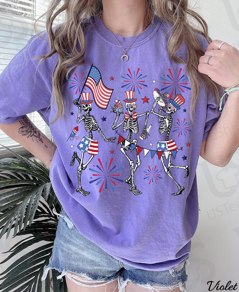 May include: A purple t-shirt featuring a festive design with three skeleton figures celebrating. They are adorned with patriotic hats and shorts, holding an American flag, cupcakes, and musical instruments. Fireworks and star accents complete the Fourth of July theme.