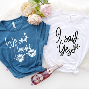 May include: Two t-shirts, one teal and one white, with text. The teal shirt says "We said Miami" with sunglasses graphic. The white shirt says "I said Yes" with a diamond graphic. A bottle of sparkling wine is in the image.