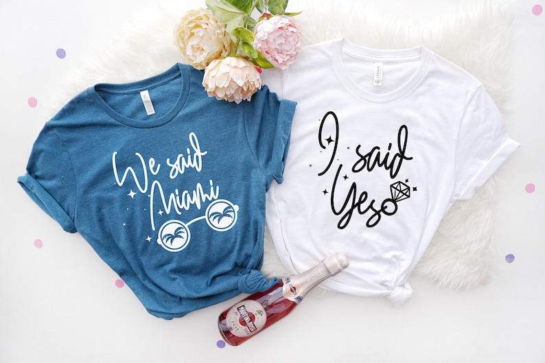May include: Two t-shirts, one teal and one white, with text. The teal shirt says "We said Miami" with sunglasses graphic. The white shirt says "I said Yes" with a diamond graphic. A bottle of sparkling wine is in the image.