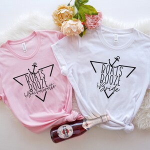 Op de afbeelding: Een lichtroze t-shirt en een wit t-shirt, elk met een zwarte afbeelding van een driehoek, een anker en de tekst "Boats Booze & Bachelorette" op het roze shirt en "Boats Booze & Bride" op het witte shirt, samen met een fles.