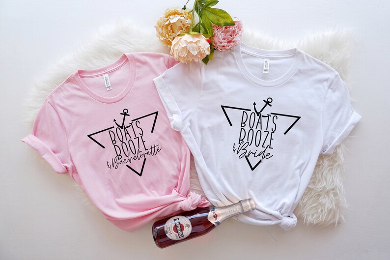 Op de afbeelding: Een lichtroze t-shirt en een wit t-shirt, elk met een zwarte afbeelding van een driehoek, een anker en de tekst "Boats Booze & Bachelorette" op het roze shirt en "Boats Booze & Bride" op het witte shirt, samen met een fles.