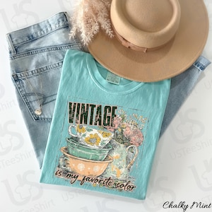 May include: A light blue t-shirt with a vintage graphic of teacups, flowers, and the text "Vintage is my favorite color." The shirt is paired with light wash jeans, a tan hat, and a necklace.
