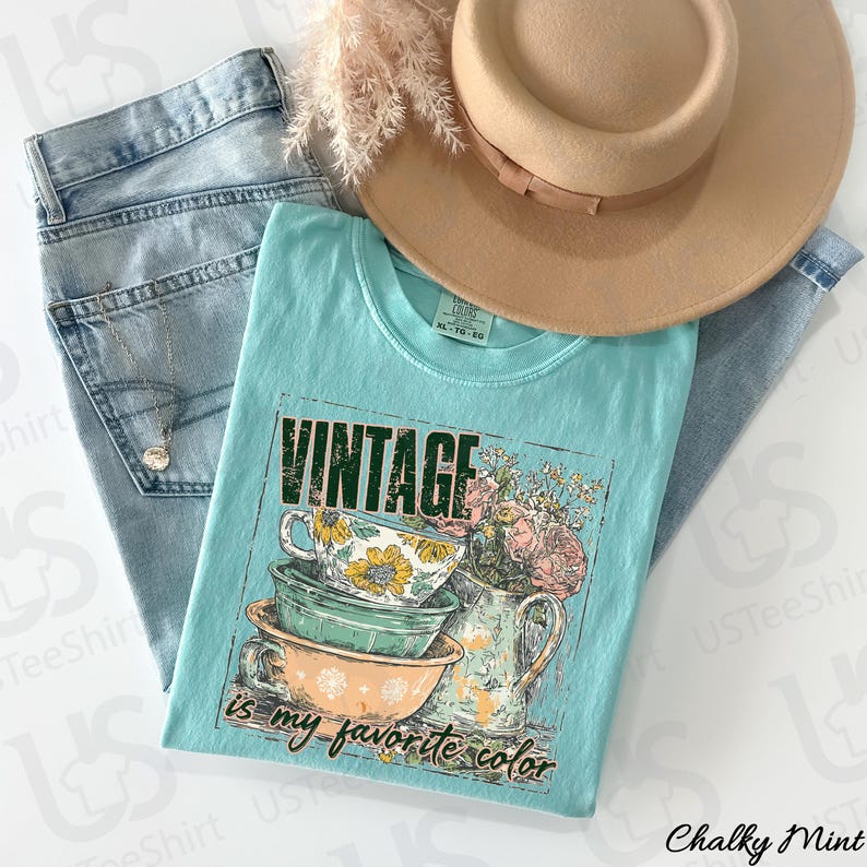 May include: A light blue t-shirt with a vintage graphic of teacups, flowers, and the text "Vintage is my favorite color." The shirt is paired with light wash jeans, a tan hat, and a necklace.
