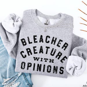 Puede incluir: Una sudadera deportiva gris con el texto "BLEACHER CREATURE WITH OPINIONS" en letras negras desgastadas. La sudadera se combina con unos vaqueros azules. Se ve un collar plateado.