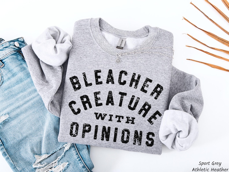 Puede incluir: Una sudadera deportiva gris con el texto "BLEACHER CREATURE WITH OPINIONS" en letras negras desgastadas. La sudadera se combina con unos vaqueros azules. Se ve un collar plateado.